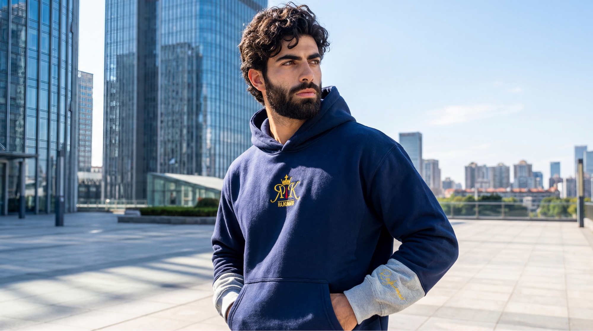 Model wearing a navy blue hoodie with a logo, standing in an urban setting with modern buildings.